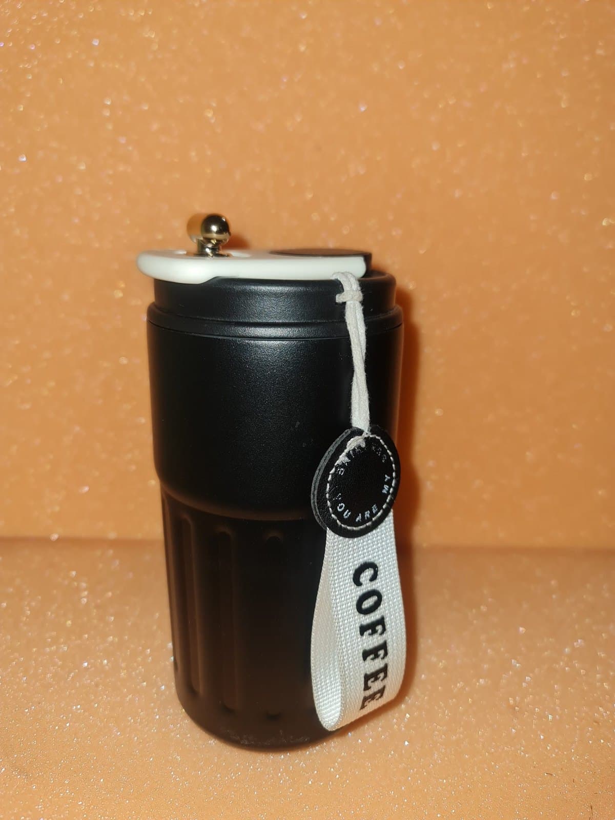 Black ribbed travel tumbler with a cream lid, gold knob, and a canvas coffee strap