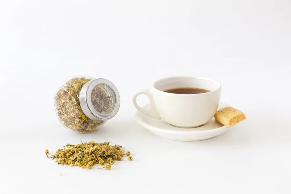 Jar of loose chamomile tea beside a white cup and saucer with a shortbread biscuit