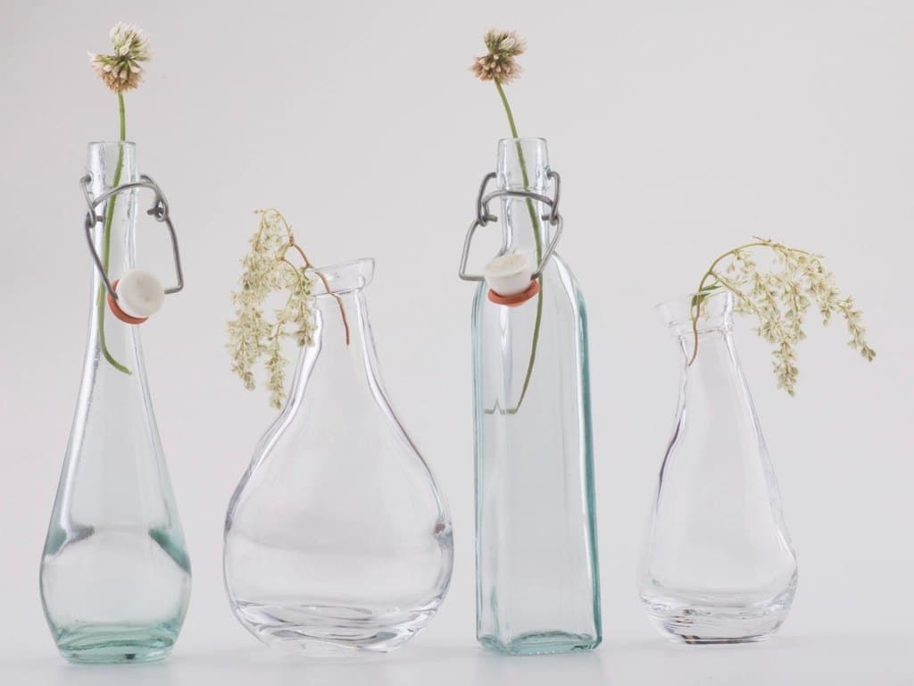 Four clear glass bud vases in mixed bottle shapes, some with swing-top closures and small white flowers