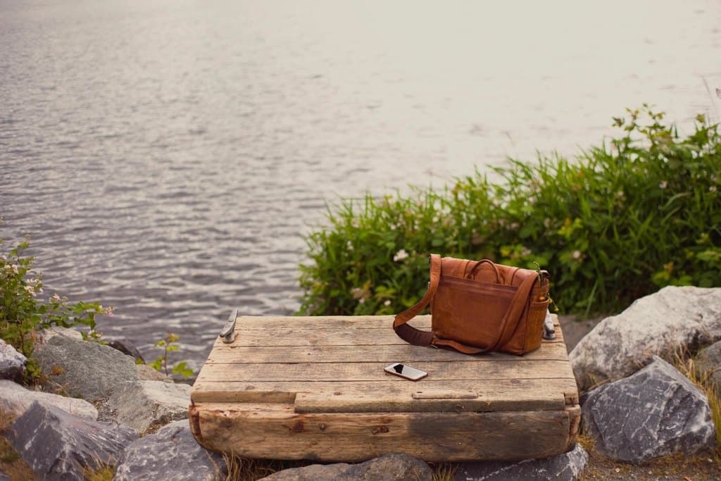 Tan leather messenger bag sitting on a weathered wooden dock beside water