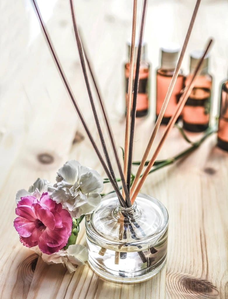Clear glass reed diffuser with rattan sticks on a pale wood table beside carnations